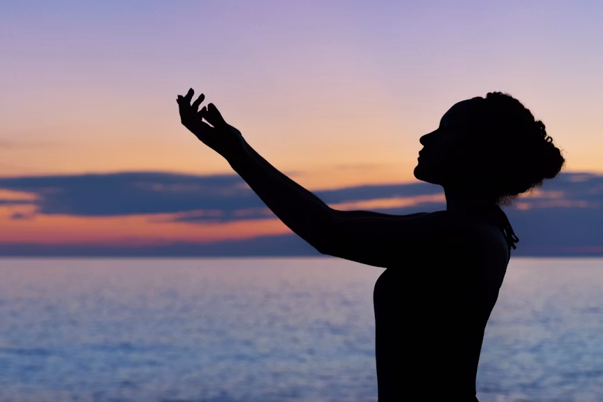 Silhouette of a woman reaching one arm upward toward a colorful sunset sky over a calm ocean.