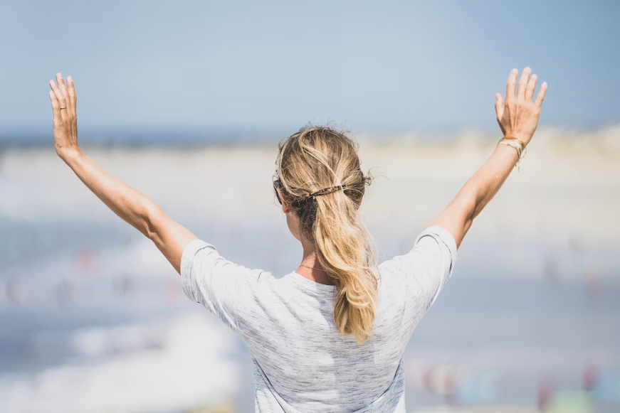 Woman with blonde ponytail in white top standing with arms raised toward a sunny beach and ocean view.