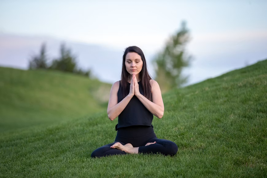 Woman with dark hair in all black sitting cross-legged in prayer pose meditating on a green grass hillside.