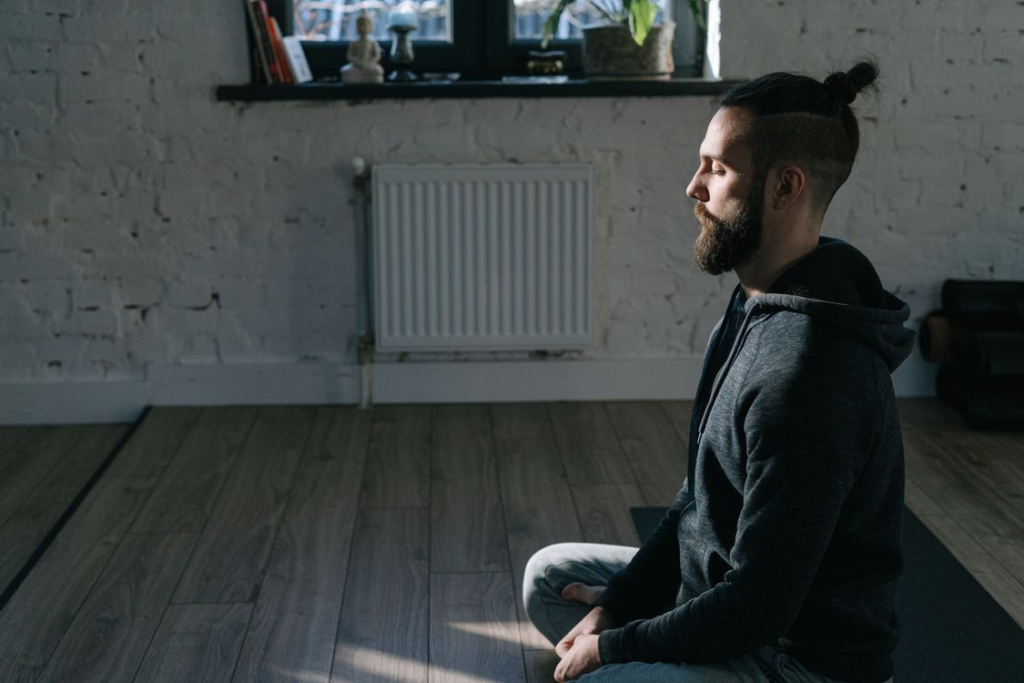 Bearded man with hair in a bun sitting cross-legged on a wooden floor meditating in a sunlit room.