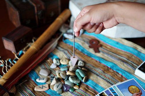 Hand holding a pendulum over a spread of rune stones and tarot cards on a colorful cloth.