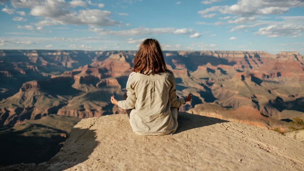 Woman in beige sitting cross-legged in meditation pose on a rocky cliff overlooking a vast canyon landscape.