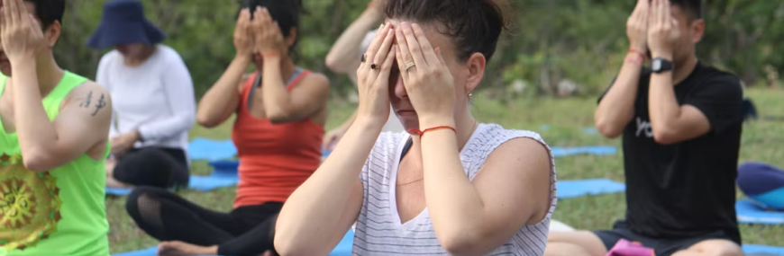 Group of people sitting cross-legged on yoga mats outdoors covering their eyes in a guided meditation exercise.