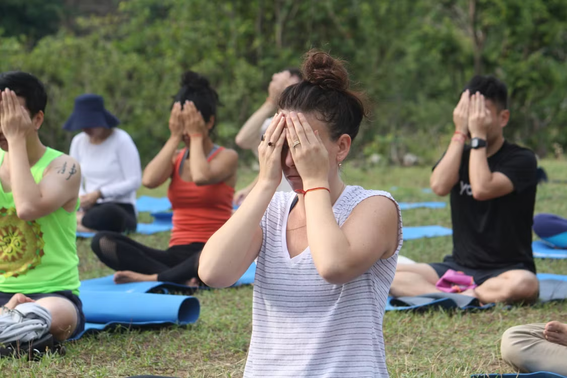 Group of people sitting cross-legged on yoga mats outdoors covering their eyes in a guided meditation exercise.
