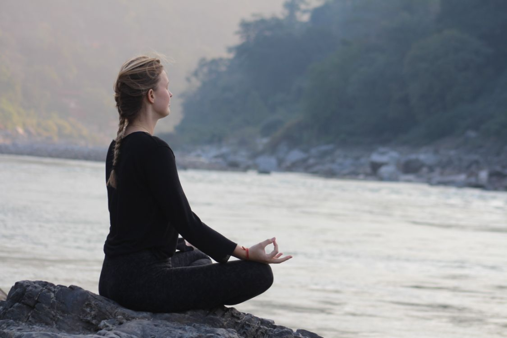 Woman with blonde braid in all black sitting cross-legged in meditation pose on rocks beside a flowing river.