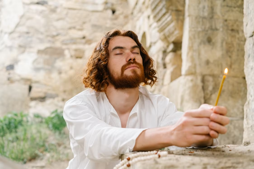 Young bearded man with curly hair in white shirt holding a lit candle with eyes closed in quiet spiritual reflection against a stone wall.