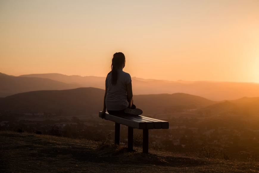 Silhouette of a woman sitting on a bench overlooking a golden sunset and mountain landscape.