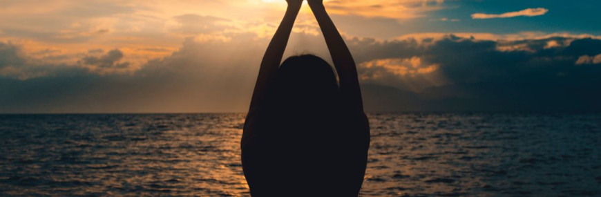Silhouette of a woman sitting by the ocean with both arms raised toward a dramatic sunset sky.