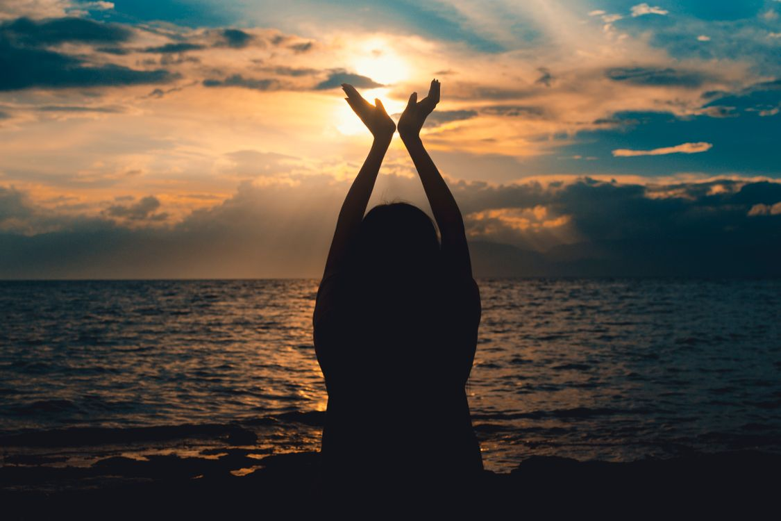 Silhouette of a woman sitting by the ocean with both arms raised toward a dramatic sunset sky.