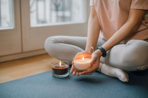 Person sitting cross-legged on a blue yoga mat holding a glowing lit candle in a peaceful indoor setting.