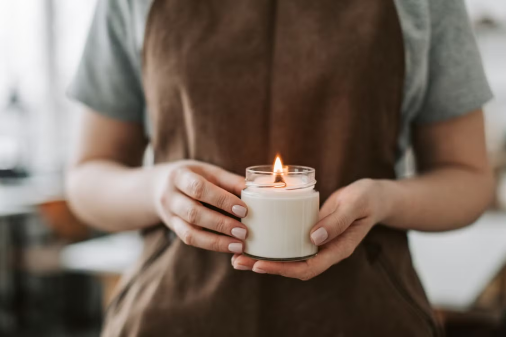 Close-up of two hands gently cradling a small lit candle in a glass jar against a soft blurred background.