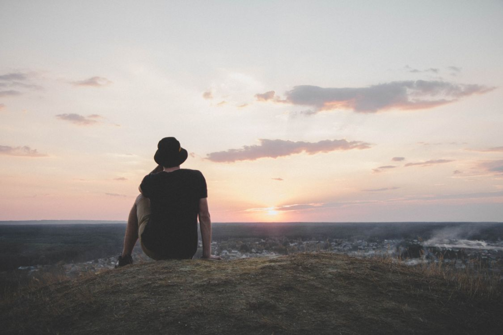 Man wearing a hat sitting alone on a hilltop overlooking a town and vast landscape at sunset.