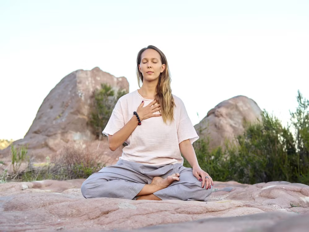 Woman with long hair sitting cross-legged on a rock with one hand on her heart and eyes closed meditating outdoors among boulders.