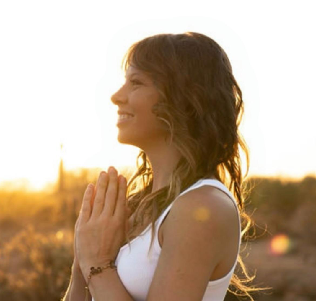 Woman standing outdoors with hands in prayer position during spiritual development practice.