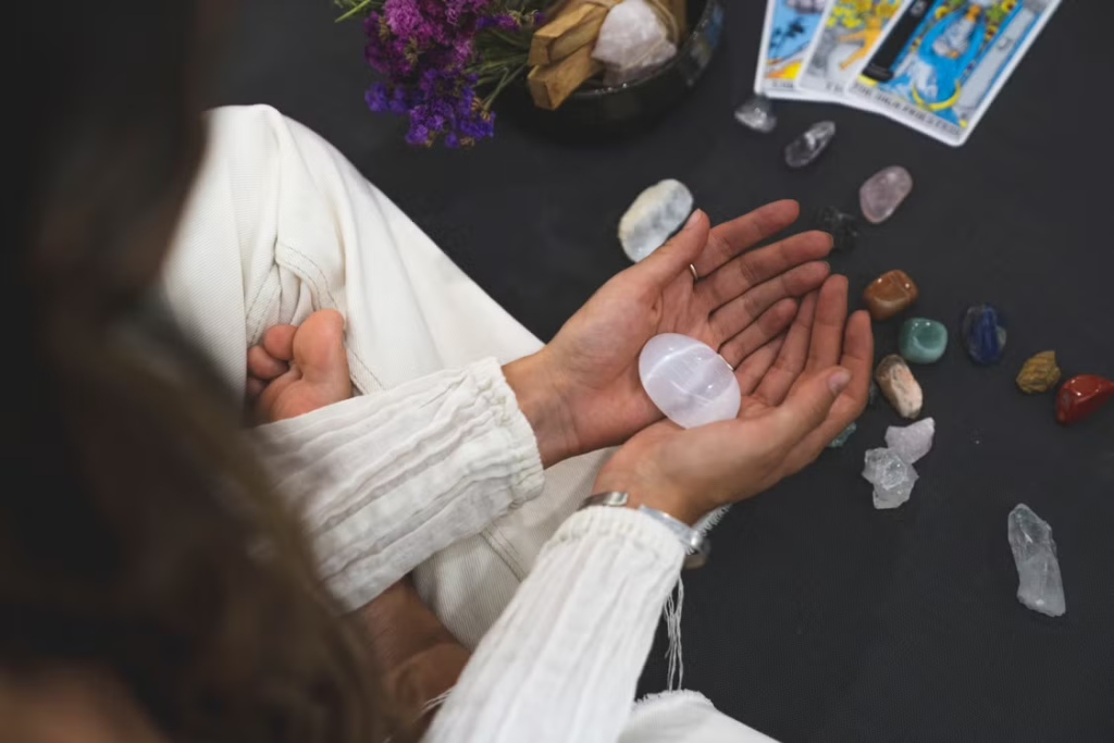 Woman in white holding a selenite crystal with colorful healing stones and tarot cards on a dark surface.