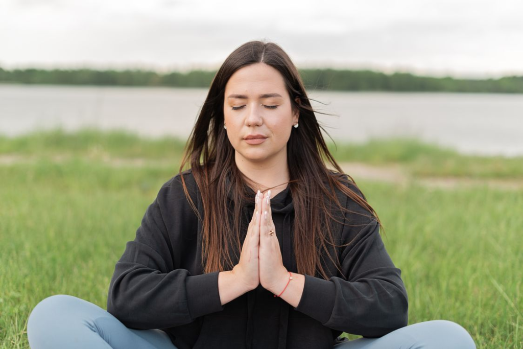 Young woman with long dark hair sitting cross-legged on grass with hands in prayer pose during outdoor meditation.