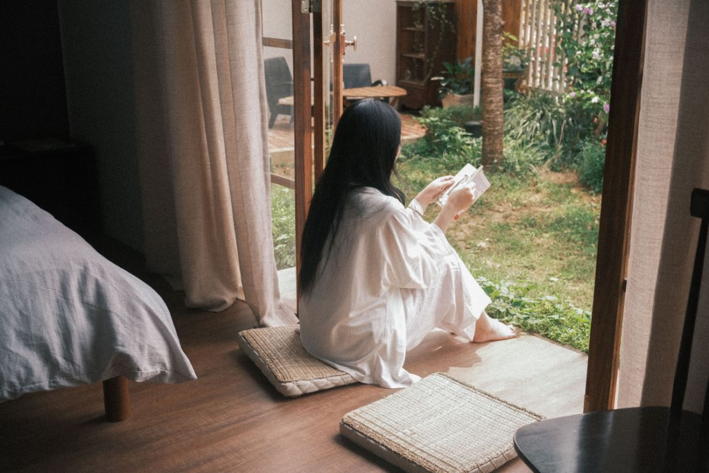 Woman with long dark hair in white clothing sitting by an open door reading a book overlooking a garden